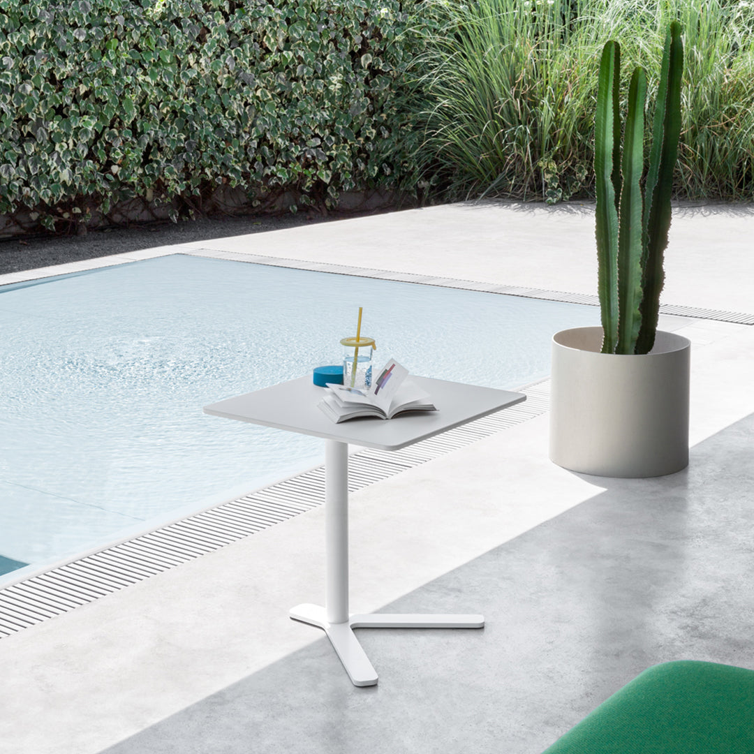 Modern white side table with a sleek gray top, set against green foliage and a poolside background.