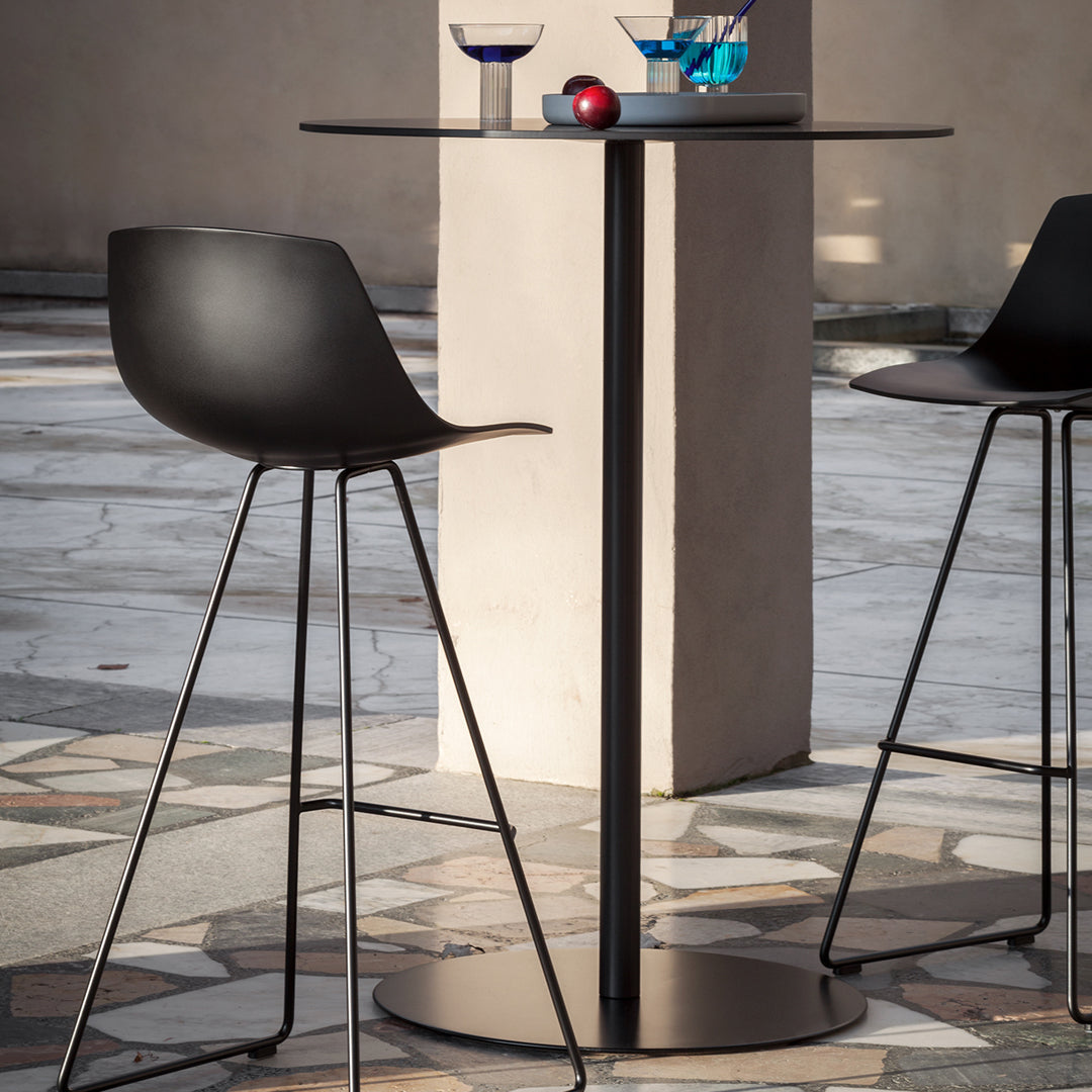 Black bar table and stools with minimalist silhouette, set against a textured stone floor pattern.