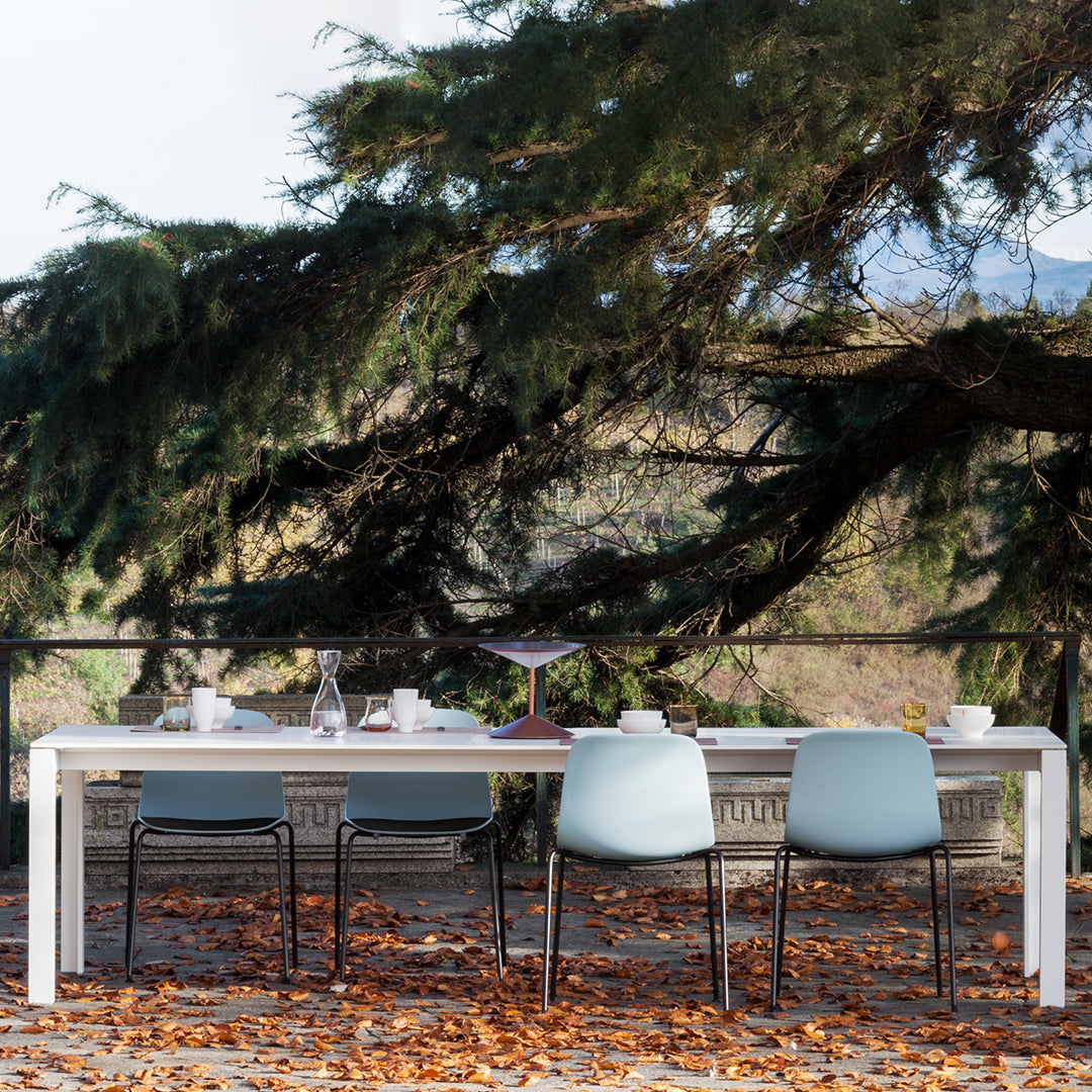 White outdoor dining table with light blue chairs, surrounded by autumn leaves and green foliage.