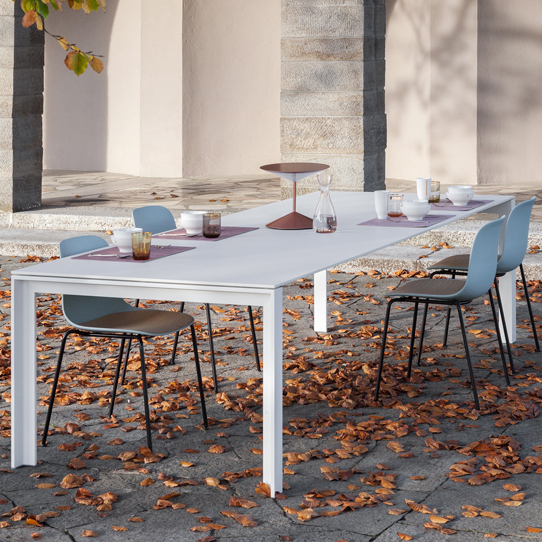White outdoor table with light blue and brown chairs, surrounded by autumn leaves on a stone patio.