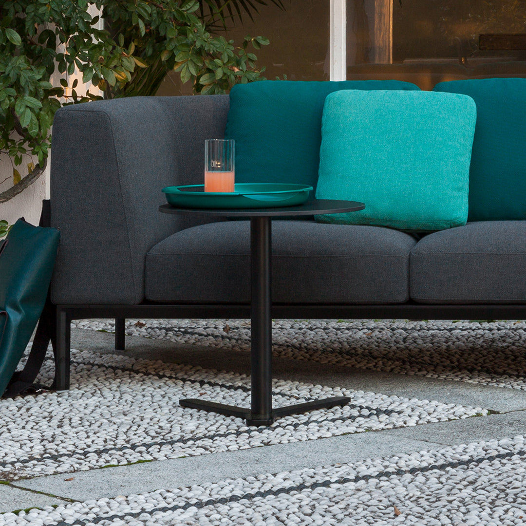Gray sofa with teal cushions and a black side table on a textured white pebble patio.