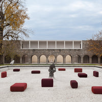 Red and burgundy outdoor cushions arranged on a gravel plaza, complemented by a soft gray sky and muted stone backdrop.