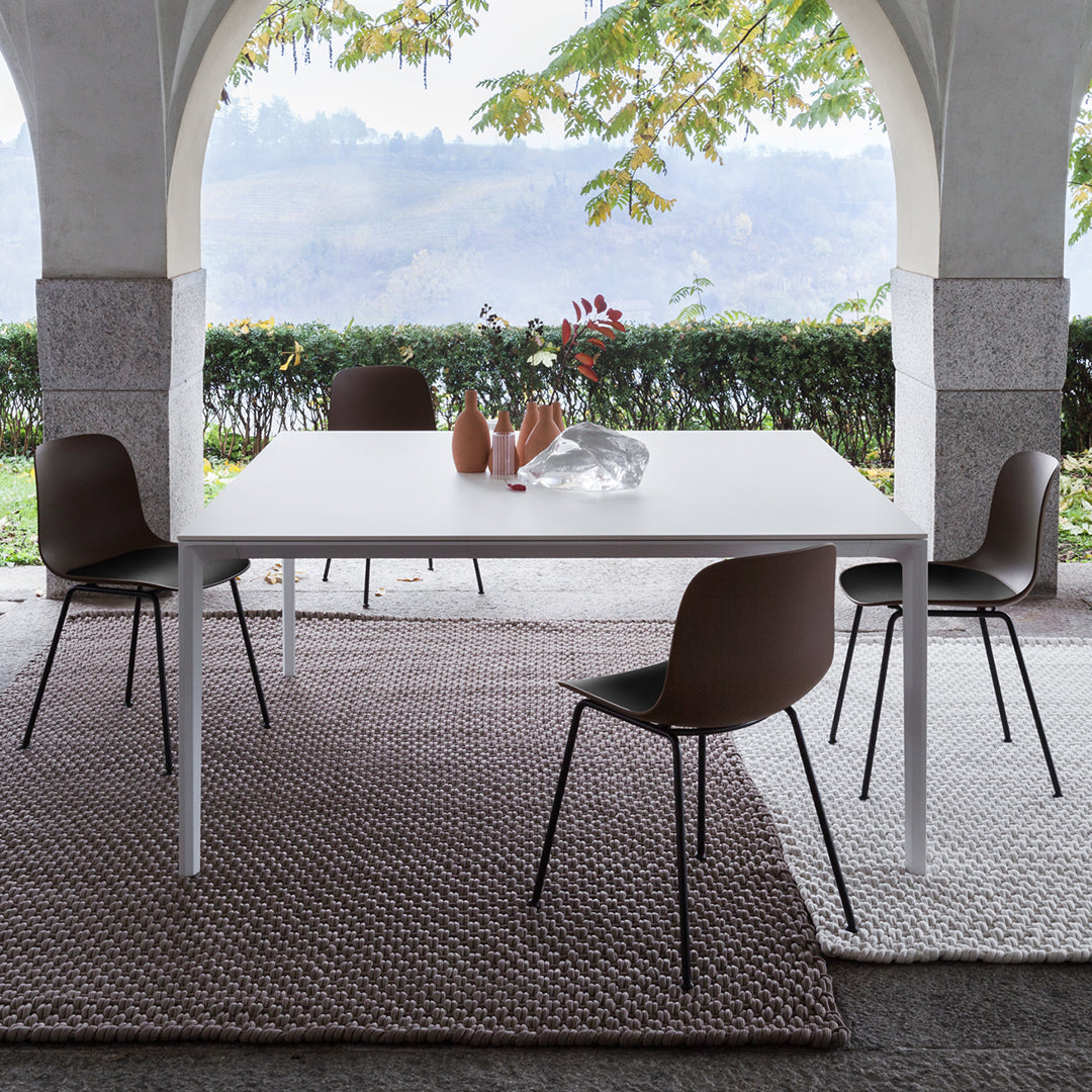 Modern outdoor dining set featuring a white table, brown chairs, and textured rugs in neutral tones. Greenery in background.