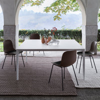 Modern outdoor dining set featuring a white table, brown chairs, and textured rugs in neutral tones. Greenery in background.