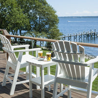 White Adirondack chairs and table on a patio, overlooking water; vibrant green grass and trees in the background.
