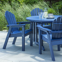 Navy blue adirondack chairs and round table set on a patio, surrounded by lush greenery and bright sunlight.