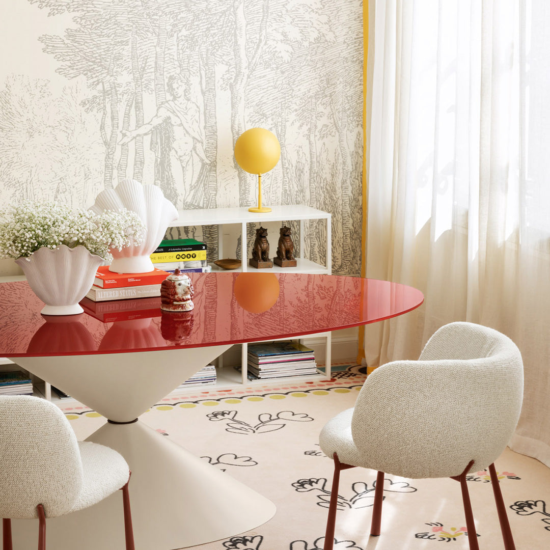 Red oval table, white upholstered chairs, yellow lamp, and patterned beige rug with floral designs against a neutral backdrop.