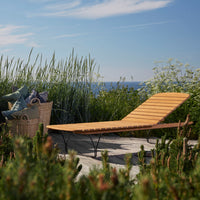 Wooden lounge chair with a sleek slatted design, set against greenery and a bright blue ocean backdrop.