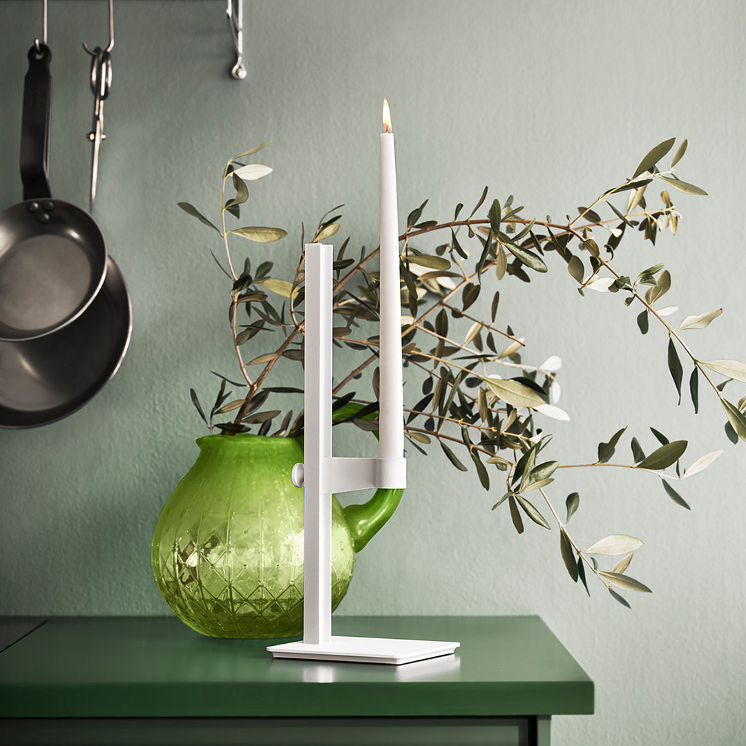 White candle holder with a tall white candle, set against a green background and a vibrant green vase with olive branches.