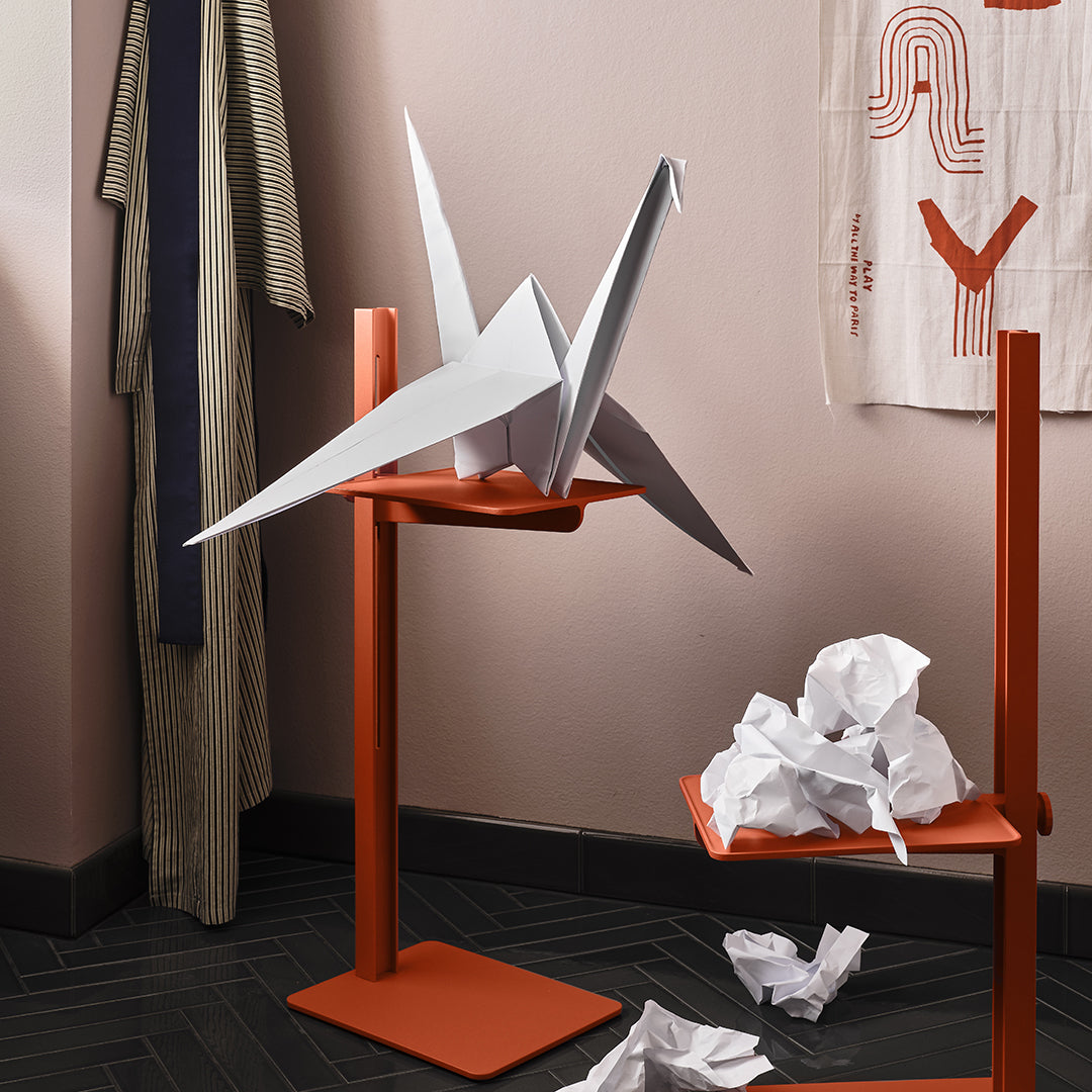 "White origami crane on orange stand with crumpled paper accents; minimalist design with soft pink wall behind."