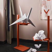 "White origami crane on orange stand with crumpled paper accents; minimalist design with soft pink wall behind."