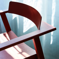 Red wooden chair with curved backrest, contrasting light wood seat, and soft blue backdrop with natural light shadows.