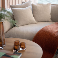 Light beige sofa with three plush pillows, complemented by a textured rust-orange throw, and a wooden coffee table.