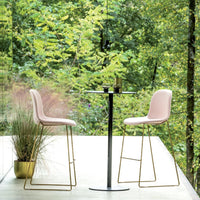 Pink upholstered bar stools with gold metal legs, set against a lush green outdoor backdrop. Minimalist design, modern style.