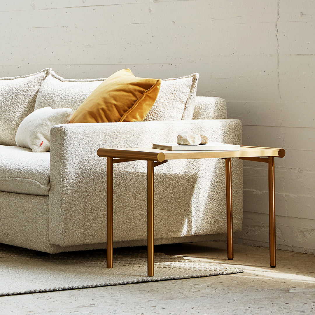 Light wooden side table with gold legs next to a white textured sofa and mustard yellow pillows, on a beige rug.