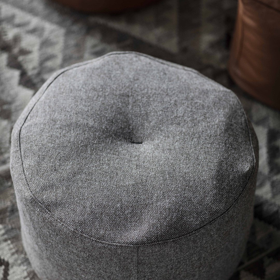 Gray round pouf with a textured finish, featuring a subtle button detail on top, set against a patterned rug backdrop.