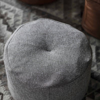 Gray round pouf with a textured finish, featuring a subtle button detail on top, set against a patterned rug backdrop.