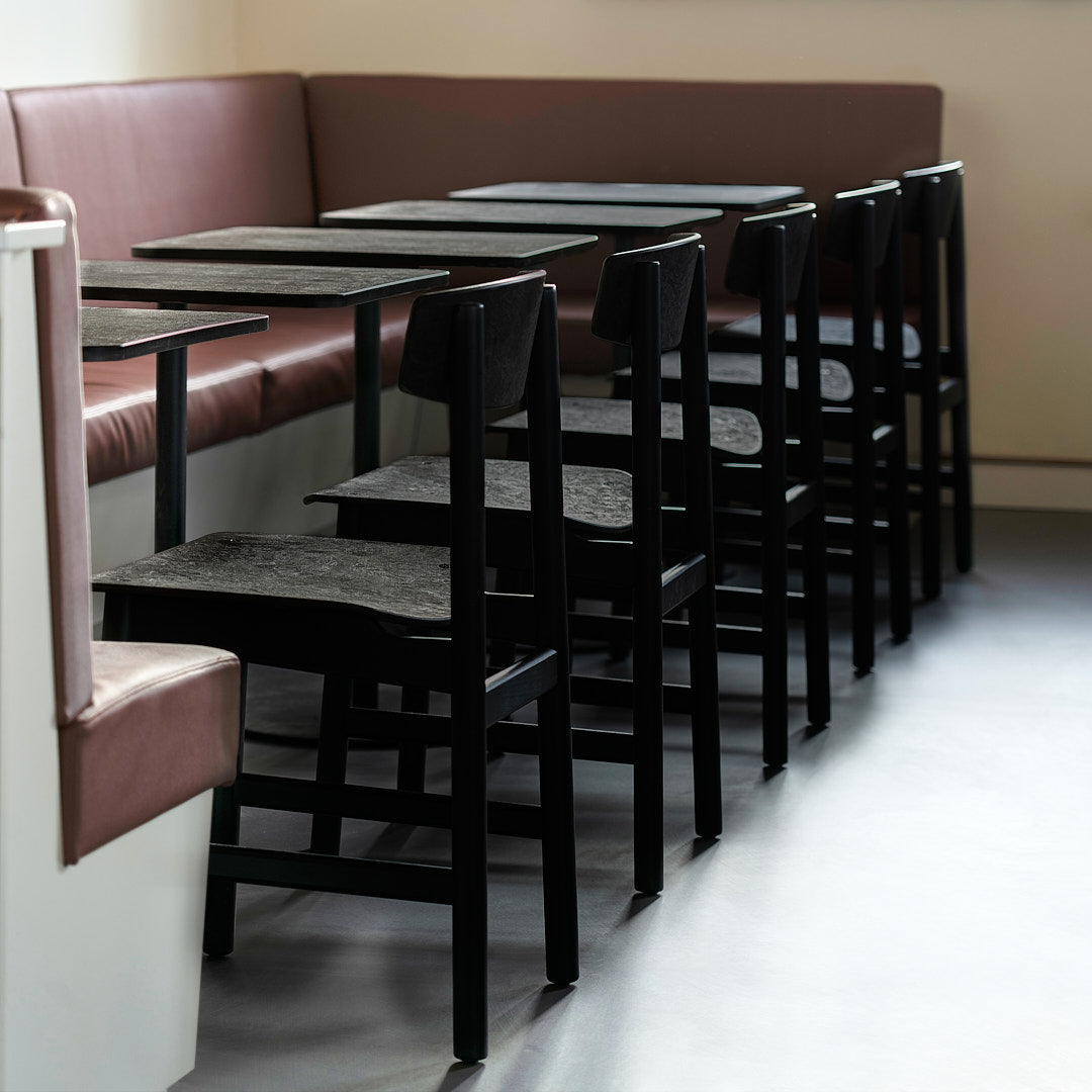 Black wooden chairs and tables with dark gray tops against a brown upholstered bench in a modern cafe setting.