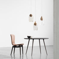 Modern wooden dining table with a round black top, paired with a brown chair and clear pendant lights. Minimalist design.
