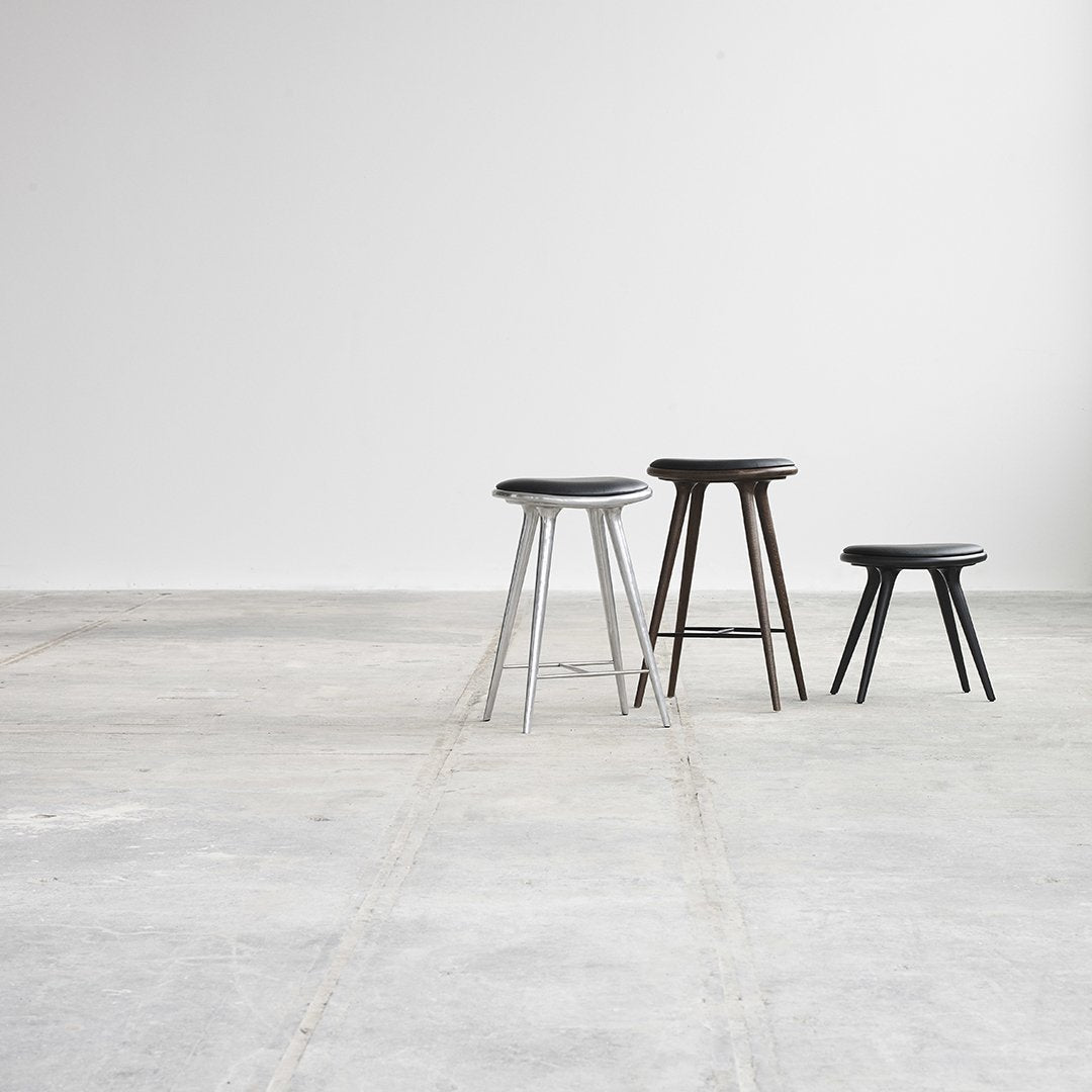 Three minimalist stools in silver, dark brown, and black on a concrete floor, showcasing modern design and clean lines.