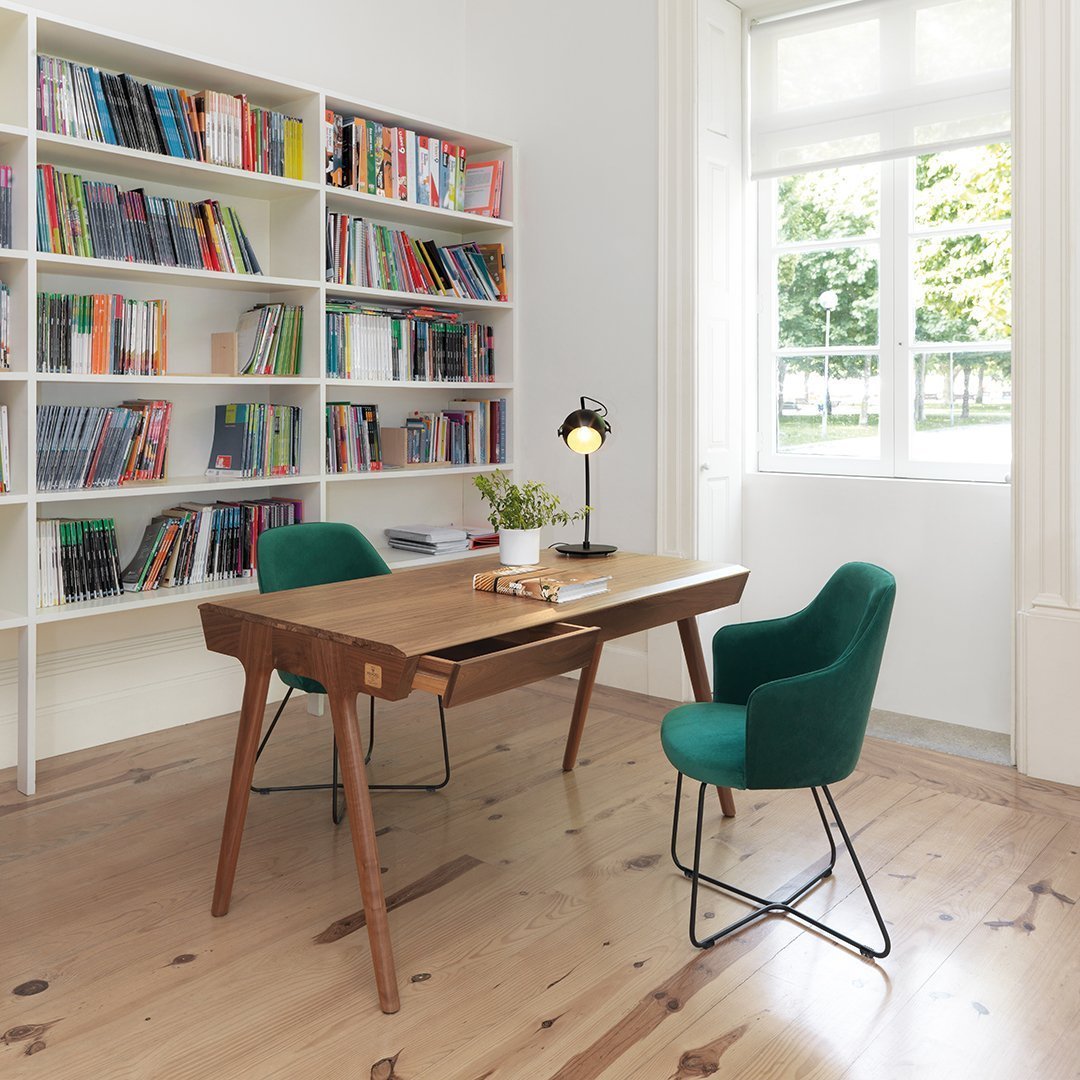Green velvet chairs with wooden desk in light-toned room; floor features warm wooden planks, bright natural light.