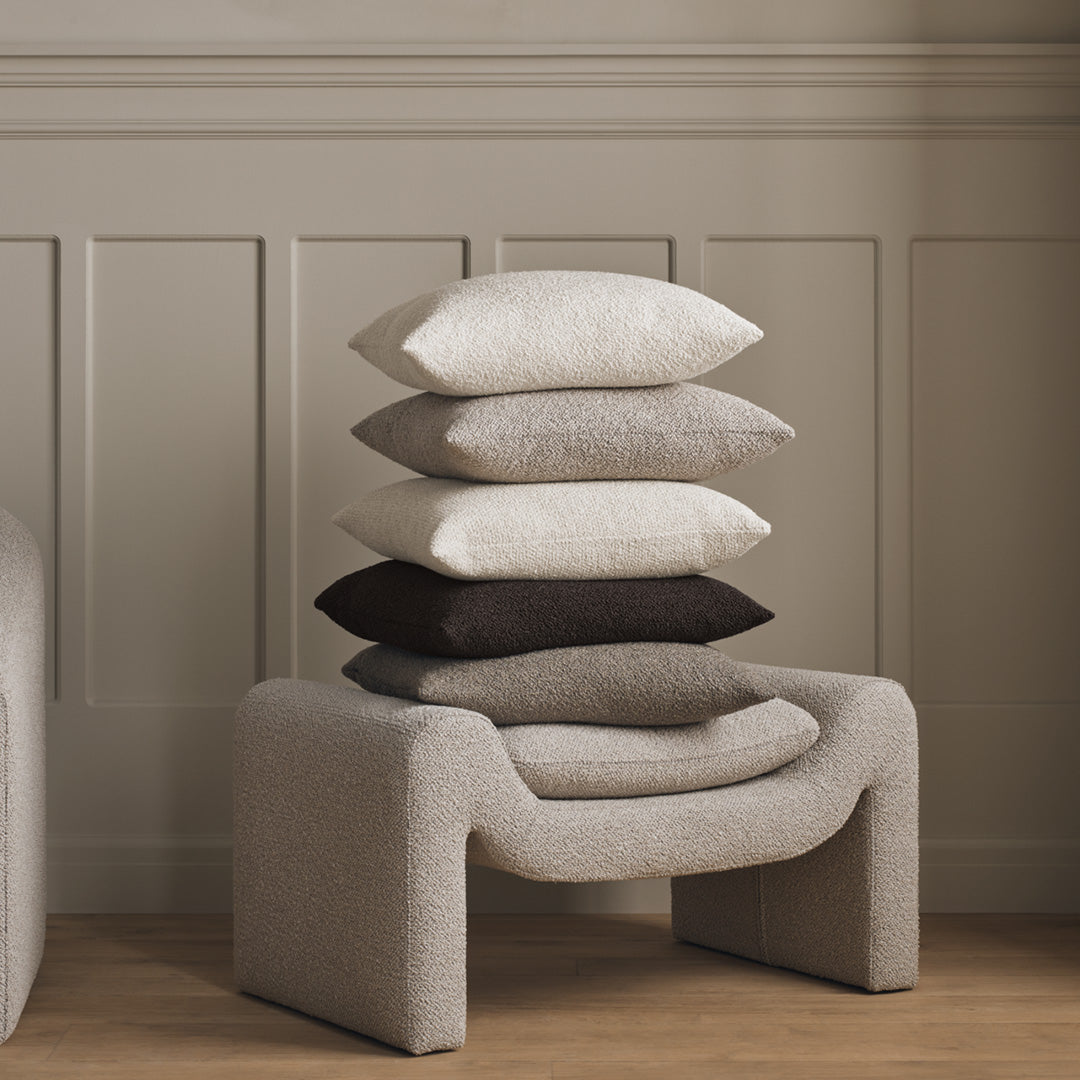 Stack of textured pillows in neutral tones: off-white, beige, gray, and dark brown, atop a modern, sculptural base.