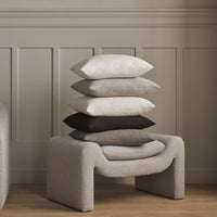Stack of textured pillows in neutral tones: off-white, beige, gray, and dark brown, atop a modern, sculptural base.