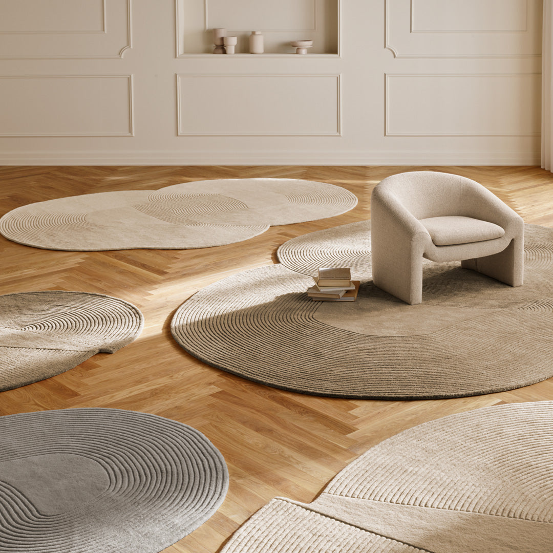 "Five round rugs in beige, gray, and cream with textured patterns on a wooden floor, complemented by a modern chair."