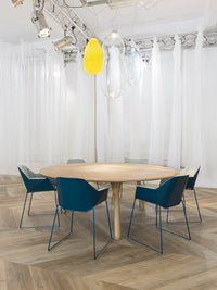 Round wooden table with blue chairs, set against a light, airy backdrop and herringbone patterned floor.