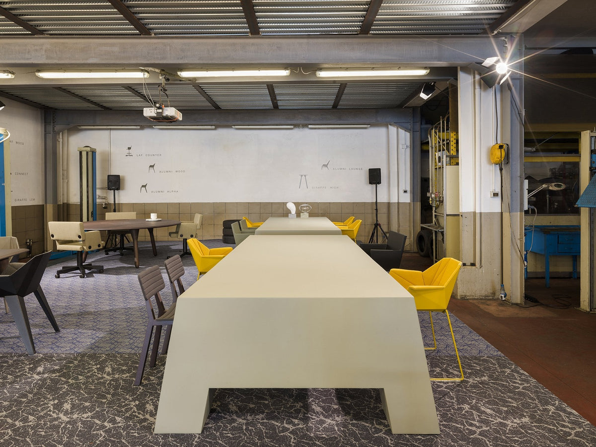 Modern workspace with a light gray table, yellow chairs, and neutral wall colors, featuring a textured floor pattern.