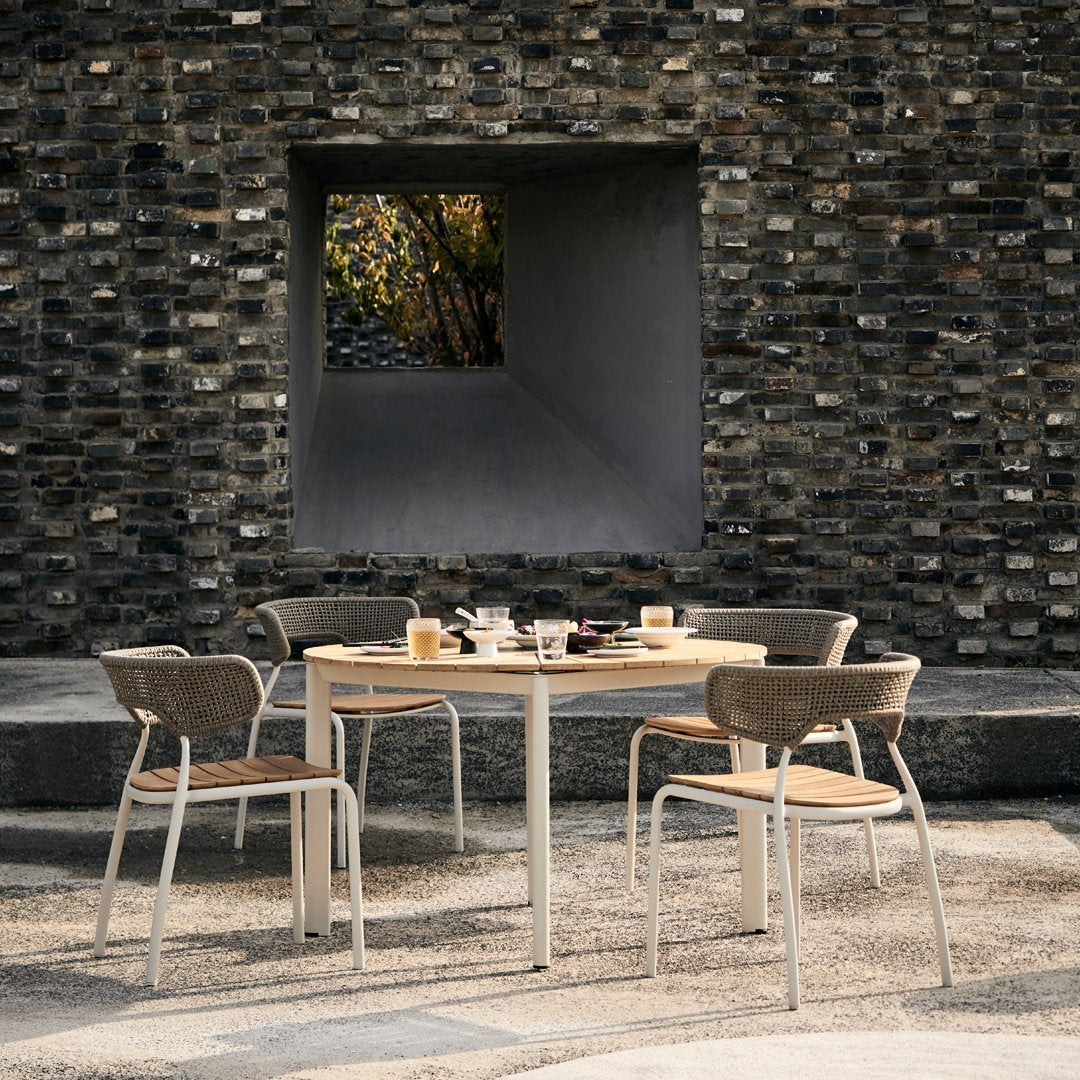 Modern outdoor dining set featuring a natural wood table and beige woven chairs against a textured gray stone wall.