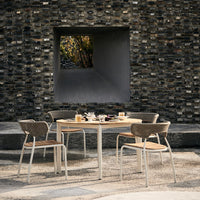 Modern outdoor dining set featuring a natural wood table and beige woven chairs against a textured gray stone wall.