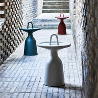 Colorful modern side tables in white, teal, and red with minimalist designs, set against a textured brick backdrop.
