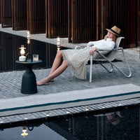 Chic white polka dot dress with beige and brown hat, relaxed by a modern black side table and serene water backdrop.
