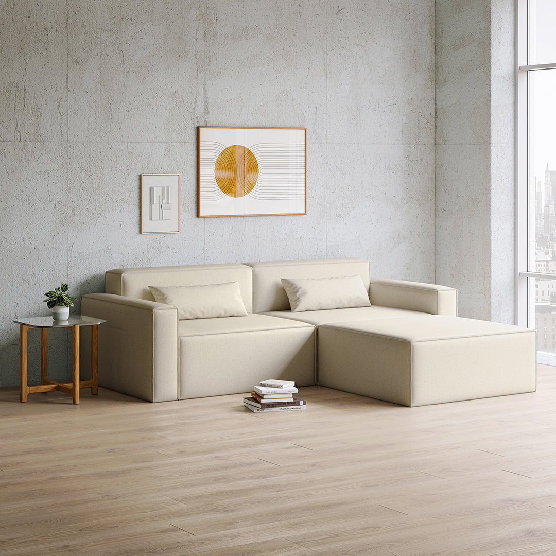 Beige modern sectional sofa in a minimalistic space with textured wooden flooring and a light gray wall.