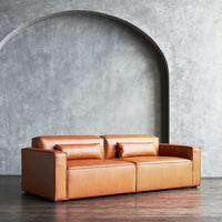 Tan leather sofa with minimalist design, featuring two matching throw pillows against a neutral gray backdrop.