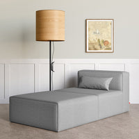 Modern gray chaise lounge with clean lines, complemented by a round wooden lamp and neutral wall backdrop.