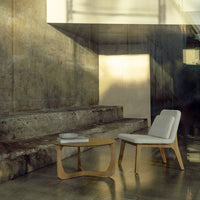 Modern white lounge chair with light wood legs and organic shapes against a textured concrete wall. Subtle shadows enhance depth.