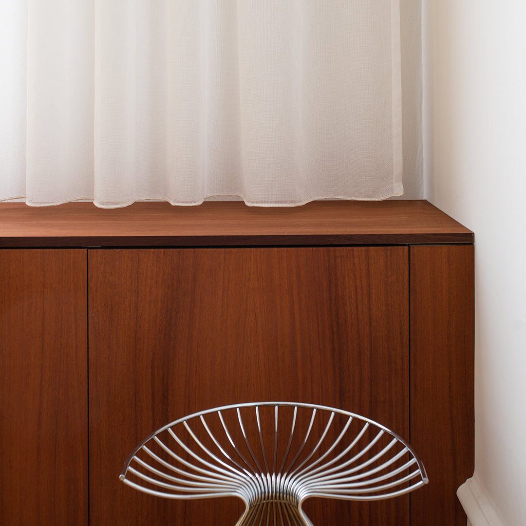 Wooden cabinet with rich brown tones, complemented by a clear, round, wire stool against soft, sheer white curtains.