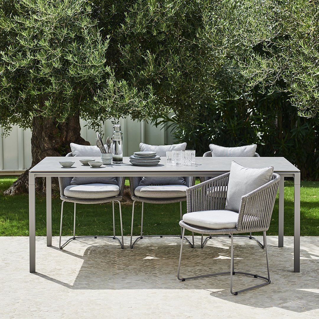 Modern outdoor dining set featuring a sleek gray table, light gray cushions, and a natural green backdrop.