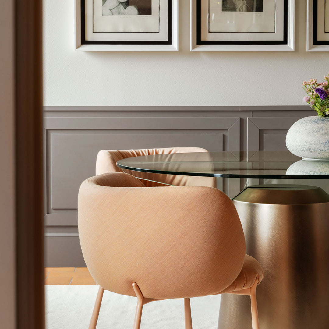 Soft pink chair with curved edges beside a glass table, complemented by neutral gray walls and framed artwork.