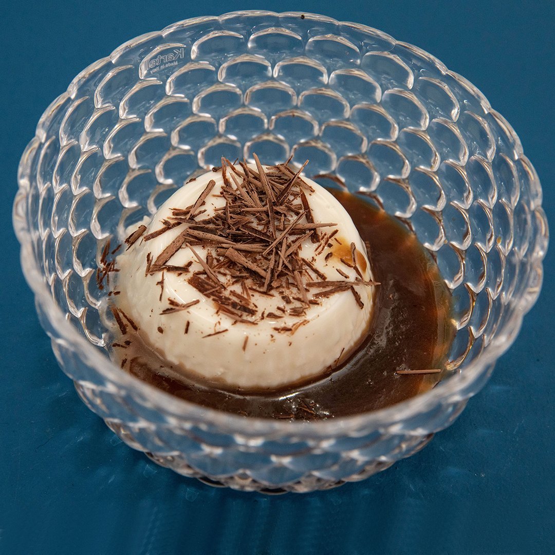 Clear glass bowl with a dotted pattern, filled with creamy dessert topped with chocolate shavings, on a vibrant blue background.
