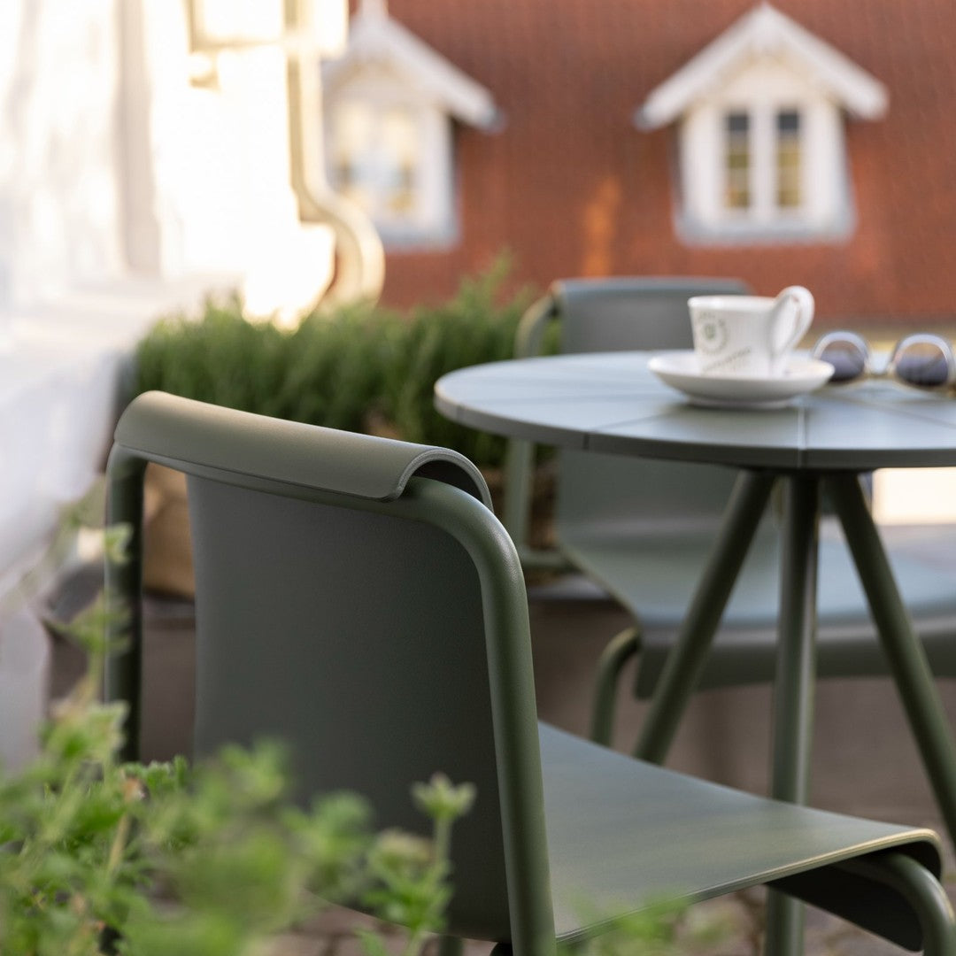 Green outdoor chair and table set with a smooth finish, surrounded by soft greenery and a warm, rustic backdrop.