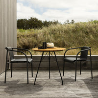 "Black metal chairs and round wooden table, set against a natural grass background under soft cloudy skies."