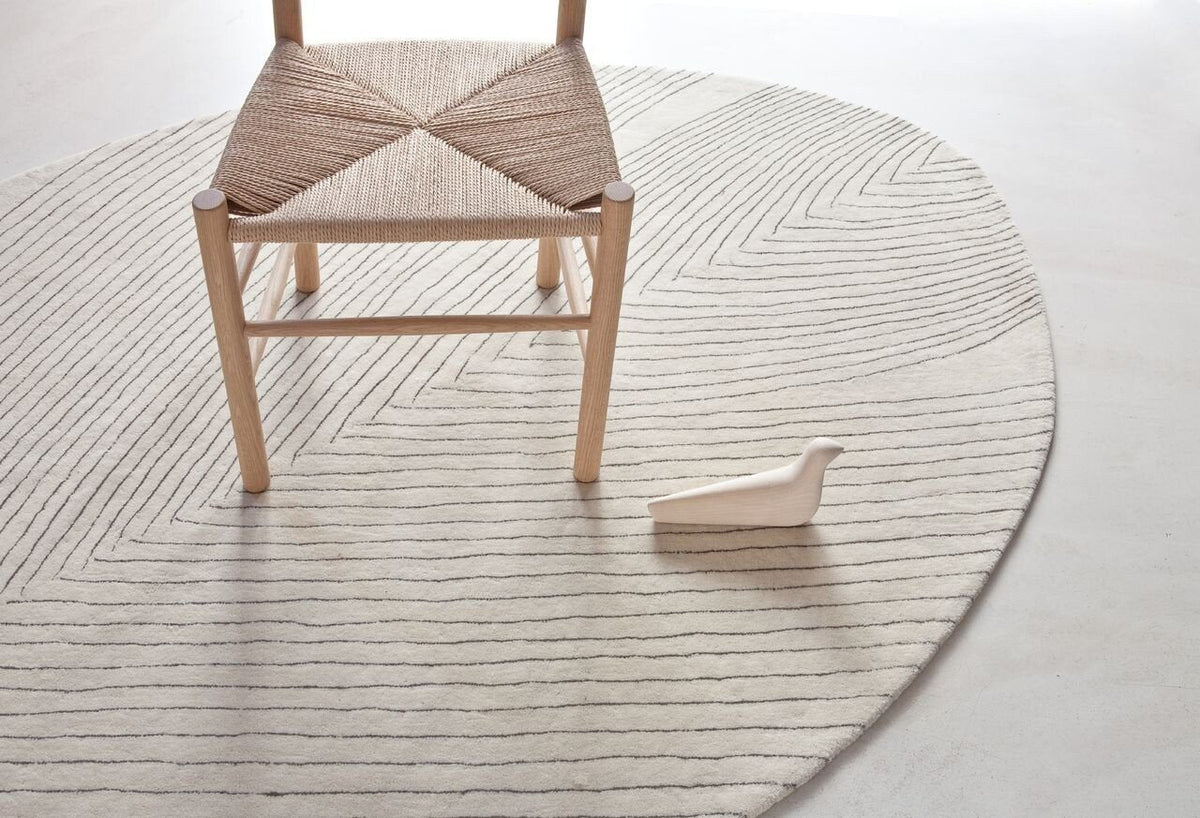 Beige geometric patterned rug with natural wood chair and light-colored decorative object. Modern, minimalist aesthetic.