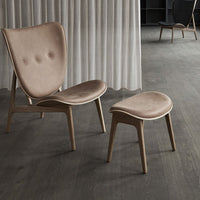 Beige velvet chair with button detailing and matching ottoman on a rustic wooden floor. Neutral tones and soft textures.