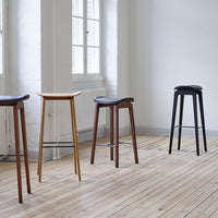 Four modern bar stools in oak, walnut, black, and natural finishes set against a minimalist backdrop.