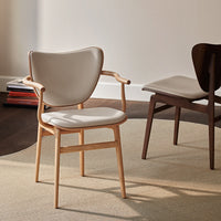 Light beige chair with a smooth finish and natural wood legs, set against a neutral-toned rug and backdrop.