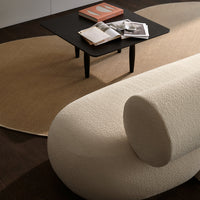 Soft white textured sofa with circular cushion, contrasted by a tan oval rug and dark wooden table. Minimalist design.