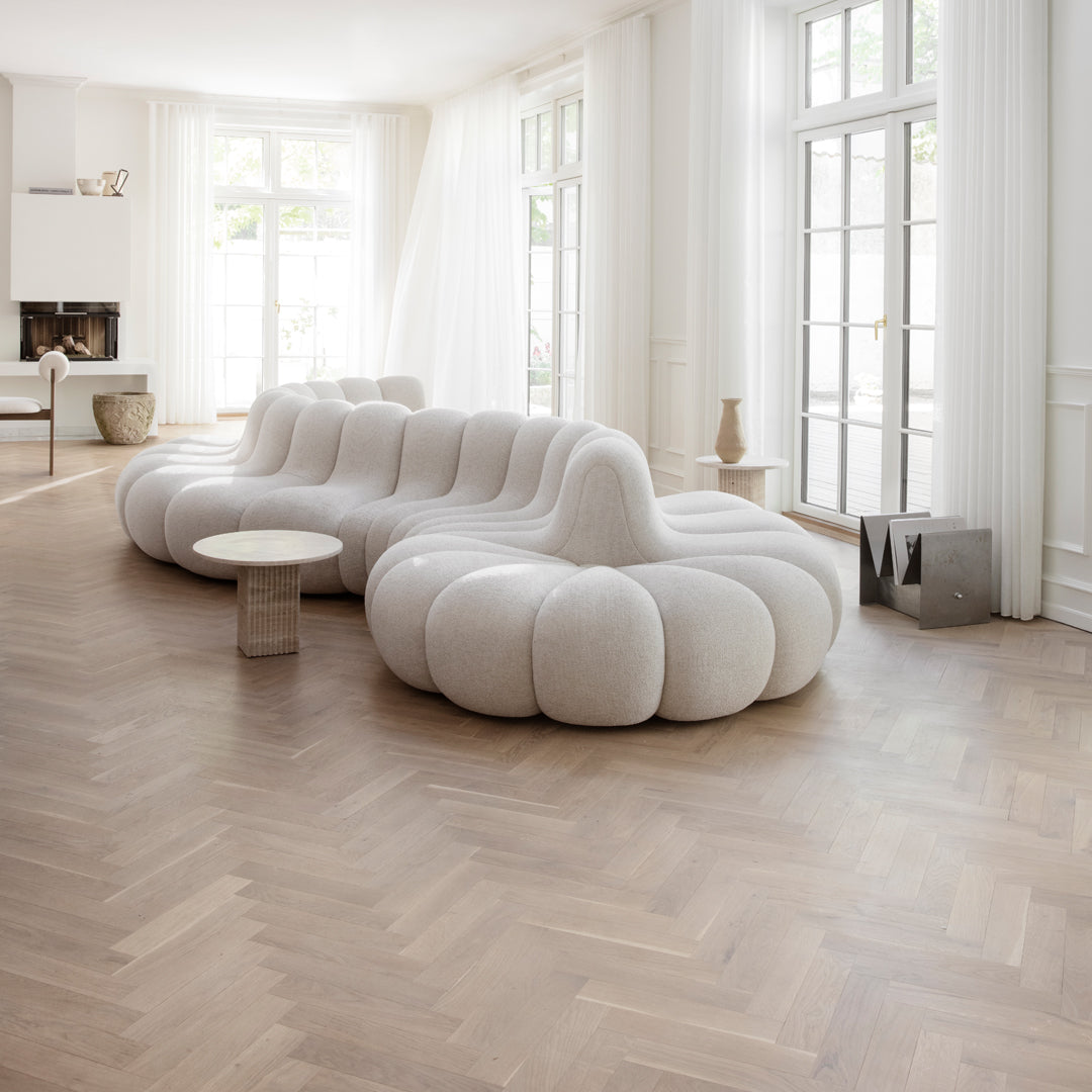 Stylish white curved sofa with soft, rounded shapes against a warm brown herringbone wood floor. Bright, airy room setting.